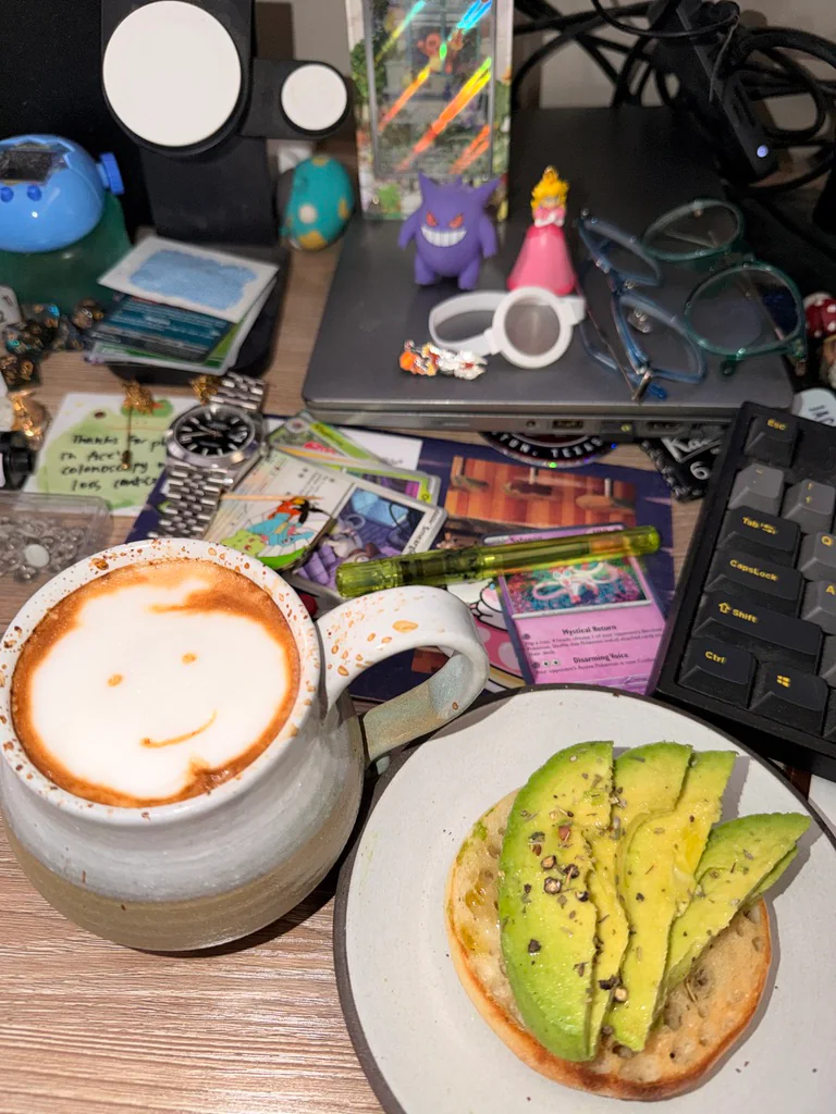 A smiling latte beside avocado toast on a desk scattered with Pokémon cards, glasses, and collectibles.