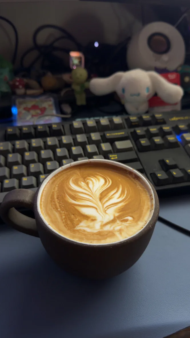 A brown ceramic cup with leaf shaped latte art on a desk beside a keyboard and a white plush toy.
