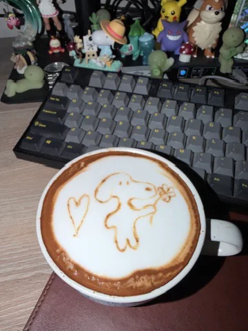 A latte with a small dog holding a flower and a heart drawn in the foam, sitting in front of a keyboard.