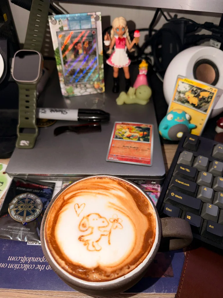 A latte with a small dog holding a flower and a heart drawn in the foam on a desk with Pokémon cards and collectibles.