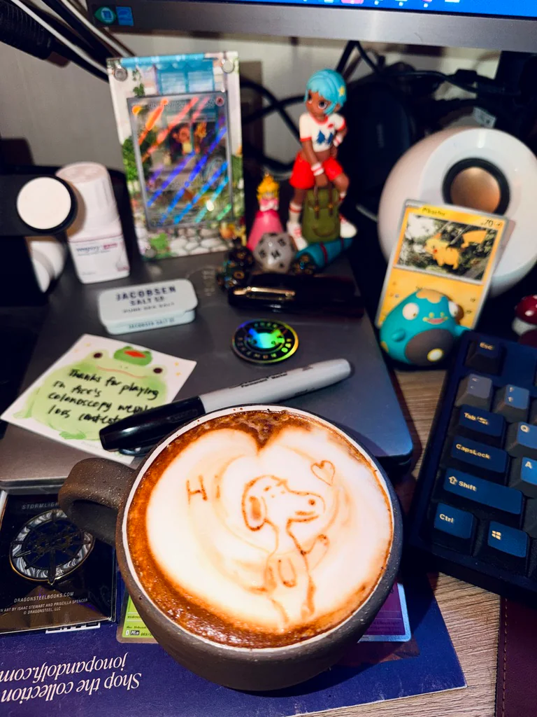 A latte with a smiling dog, a heart, and the word Hi drawn in the foam on a desk with Pokémon collectibles.