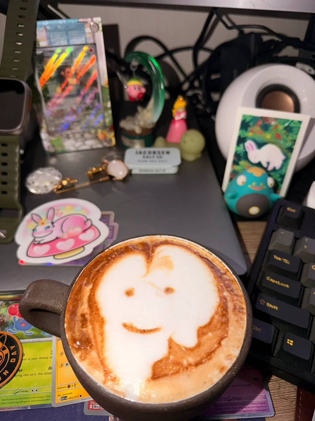 A smiling ghostlike face in the foam of a latte on a desk with Pokémon collectibles.