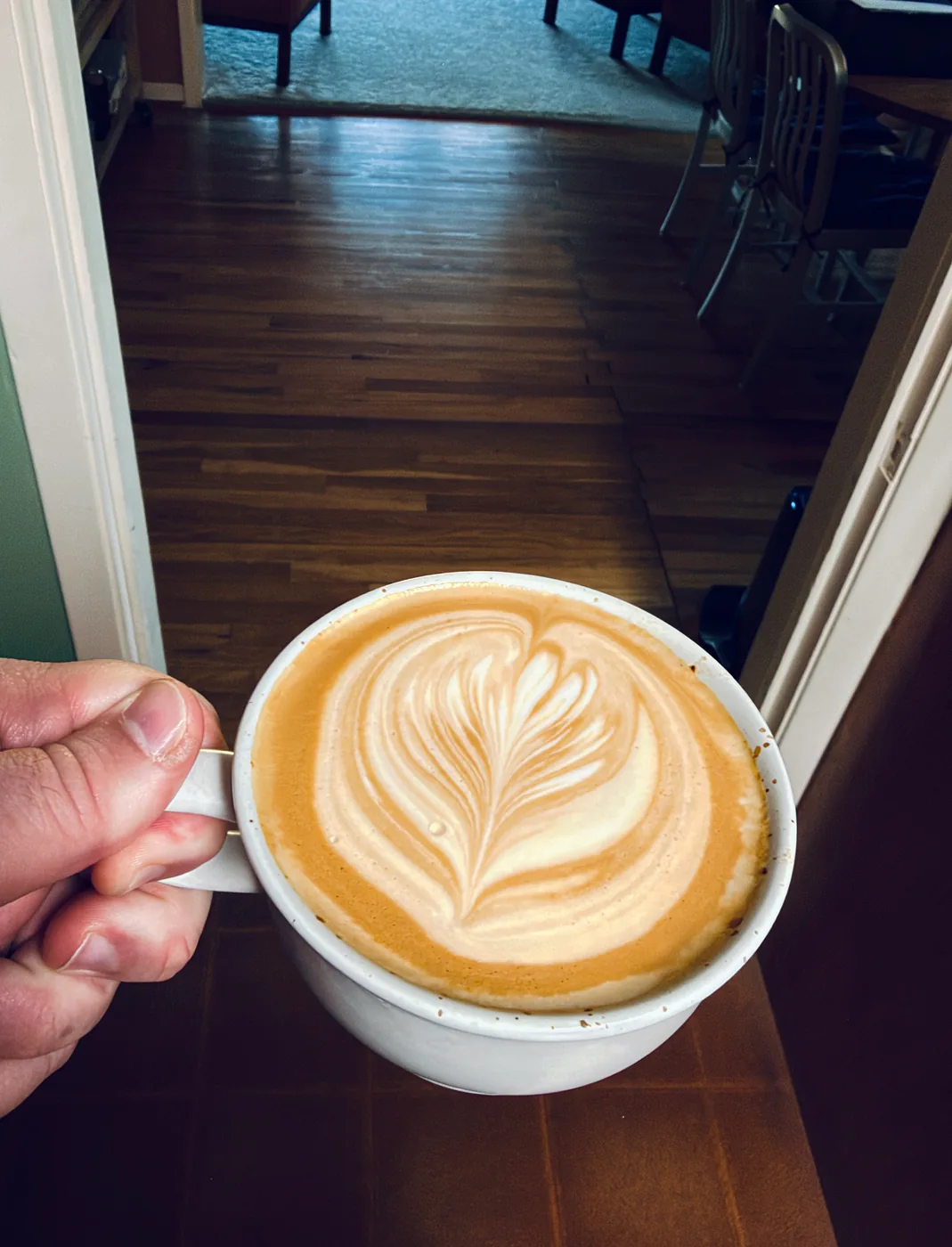 A hand holding a white cup with a finely layered leaf poured into the latte.