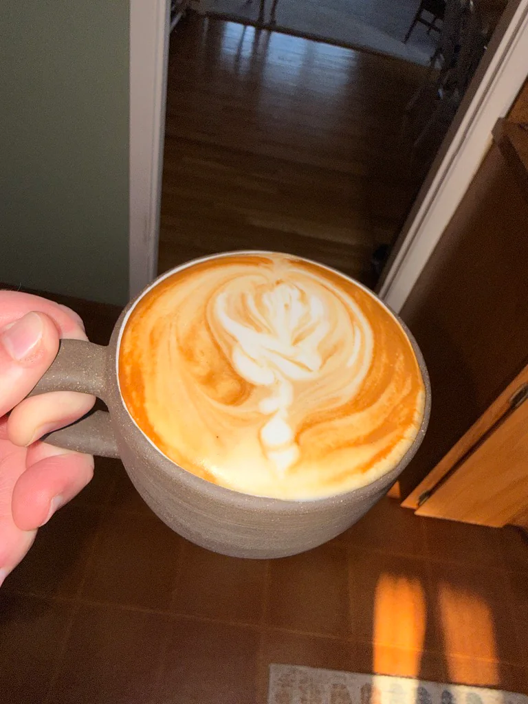 A hand holding a gray ceramic cup with a leaf pattern poured into the latte foam.