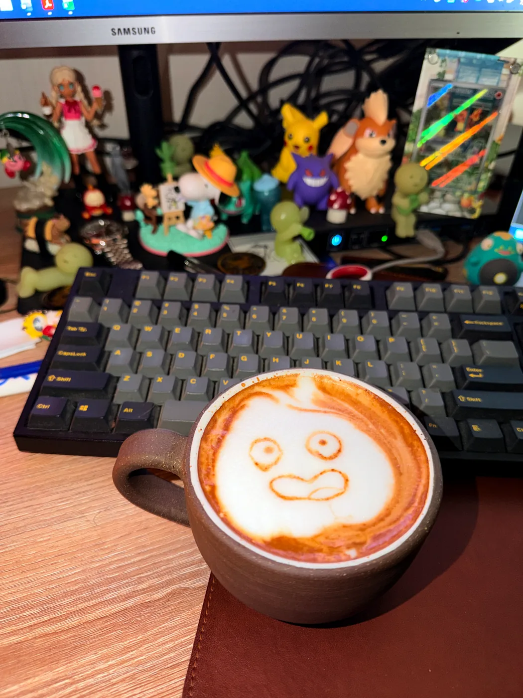 A wide eyed, surprised face drawn in latte foam on a desk with Pokémon figures.