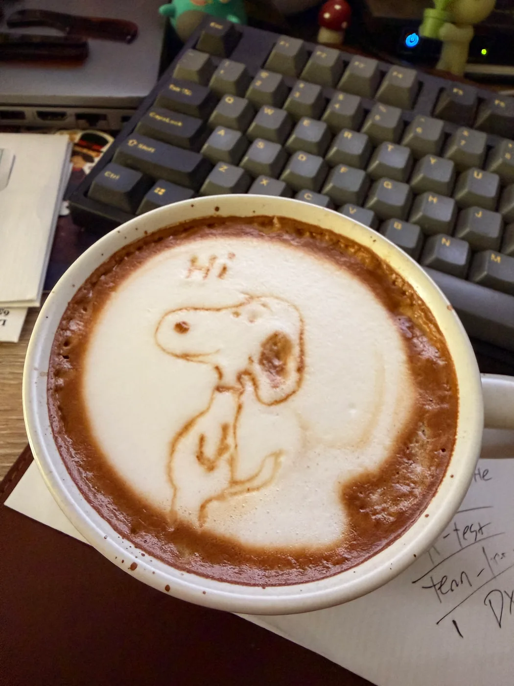 A Snoopy drawing with the word Hi in the foam of a latte on a desk.