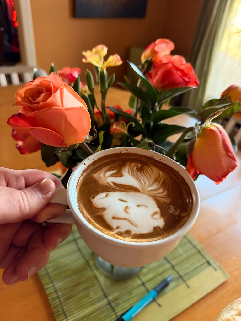 A latte with an animal face drawn in the foam, held in front of a bouquet of coral roses.
